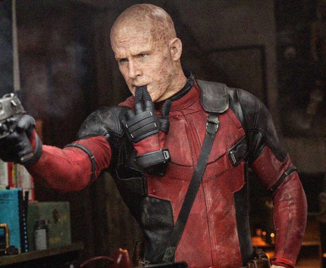 A person in a red and black superhero costume points a gun while putting their finger to their mouth. Their face has a scarred appearance, and they wear a serious expression in a dimly lit indoor setting.