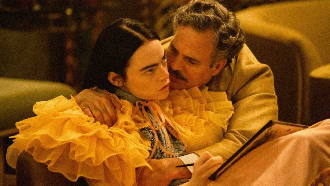 A man in a tan suit hugs a woman in a ruffled yellow dress. The woman looks away with a serious expression while the man leans in closely, his arm around her shoulders. The scene appears intimate and emotional.