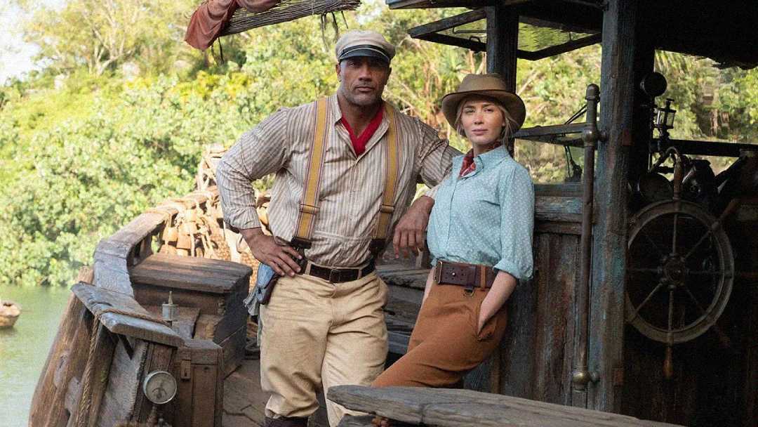 Two people dressed in early 20th-century adventure clothing stand on a rustic wooden boat, surrounded by lush greenery. One wears suspenders and a cap; the other wears a hat, scarf, and blouse.