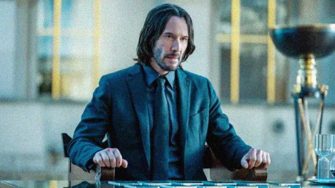 A man with shoulder-length dark hair and a beard, dressed in a dark suit and tie, sits at a table with a focused expression. The background is blurred with large windows and metallic objects visible.