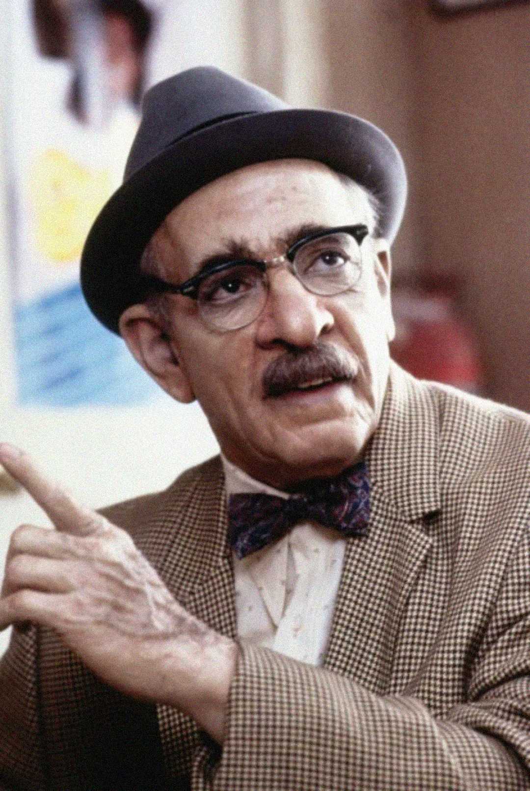 An older man with a mustache, wearing round glasses, a gray hat, a bow tie, and a checked jacket, gestures with one hand while looking to the side.