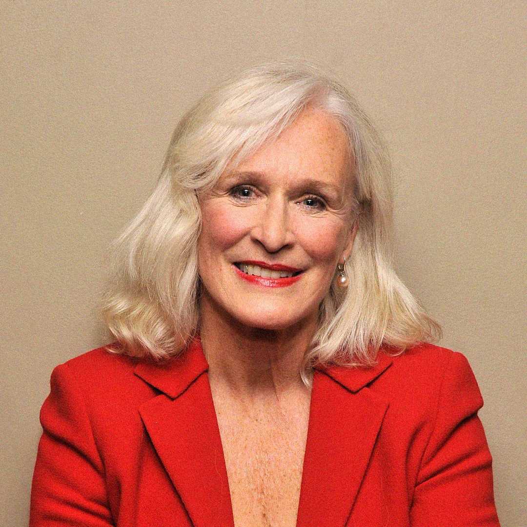 An older woman with shoulder-length white hair smiles at the camera. She is wearing a red blazer and red lipstick, posed against a plain beige background.