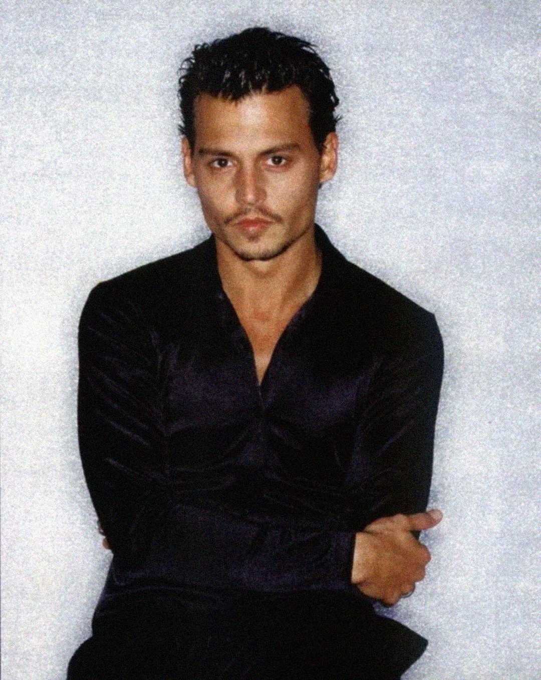 A man with short, slicked-back dark hair and a mustache is wearing a black shirt with sleeves rolled up, sitting against a light gray background with his arms crossed.