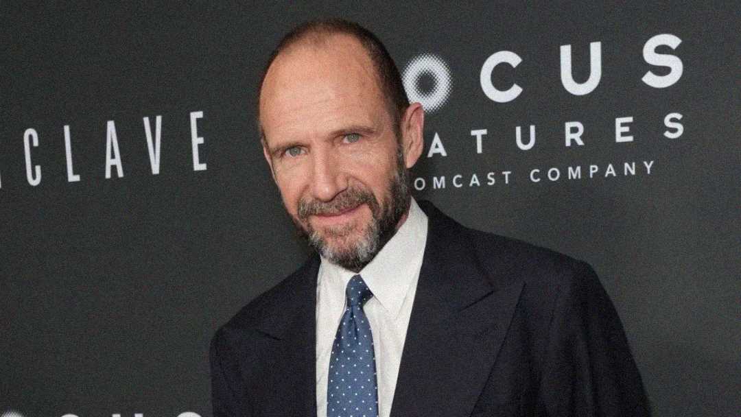 A man in a dark suit, white shirt, and tie stands in front of a backdrop with the words "Focus Features" and "Enclave." He has a trimmed beard and is looking at the camera with a slight smile.