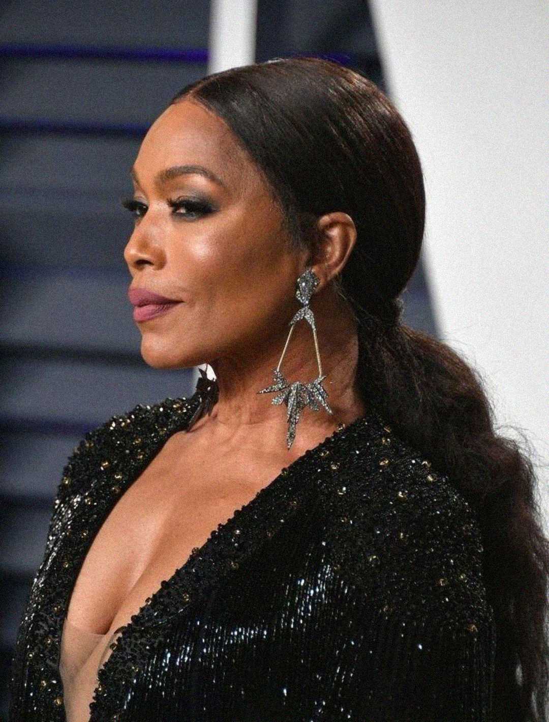A woman in a black, sparkling dress with a deep neckline poses at an event. She has long, dark hair styled in a low ponytail and wears large, dangling earrings shaped like stars. The background is blurred and dark-toned.