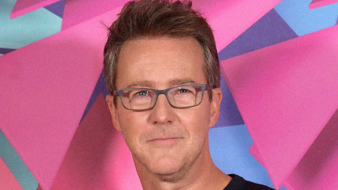 A man with short brown hair and glasses is smiling slightly, standing in front of a colorful, abstract pink and blue background.