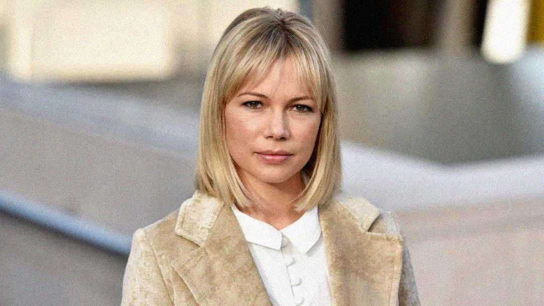 A woman with straight blonde hair and bangs, wearing a light-colored coat over a white shirt, stands outdoors in soft natural light, looking directly at the camera with a neutral expression.