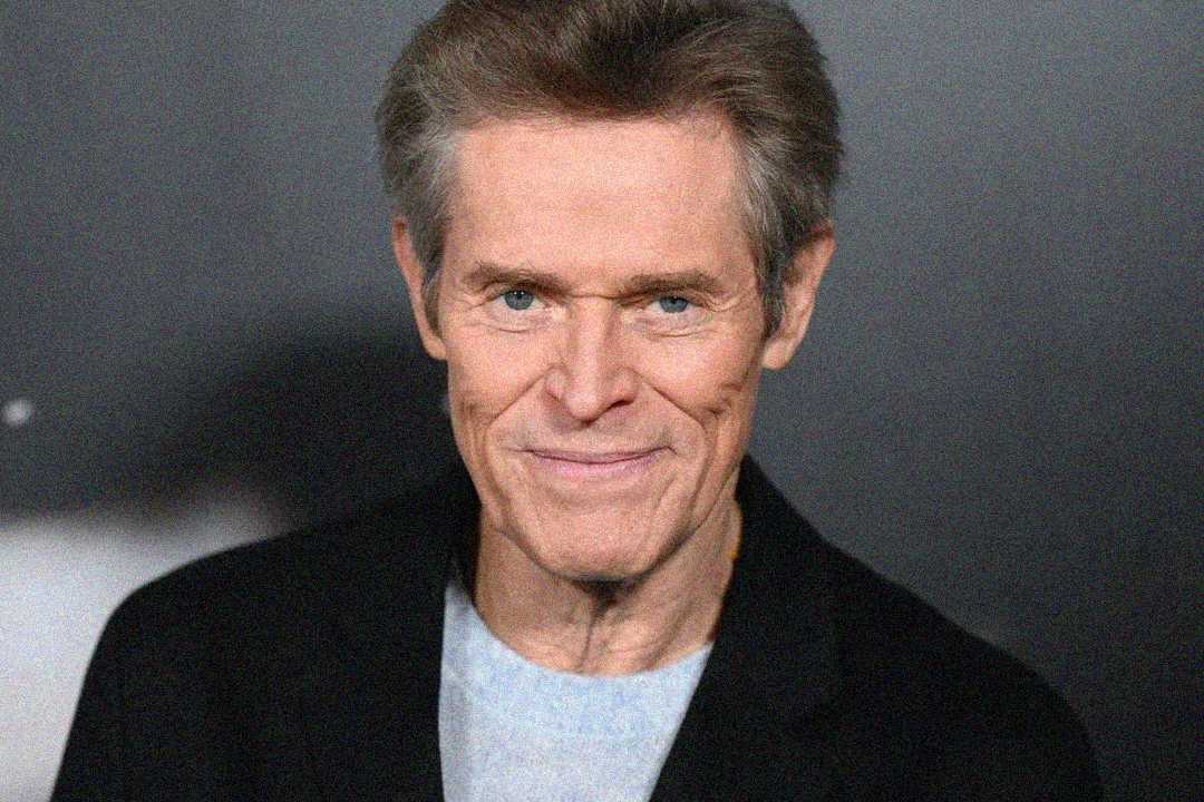A middle-aged man with light skin, short brown hair, and blue eyes smiles slightly while wearing a black jacket over a gray shirt against a dark blurred background.