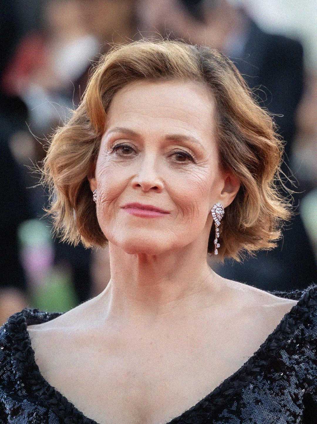 A woman with short, wavy light brown hair and diamond earrings is wearing a dark, sparkly off-the-shoulder dress. She is posing and smiling slightly at a formal event with a blurred crowd in the background.