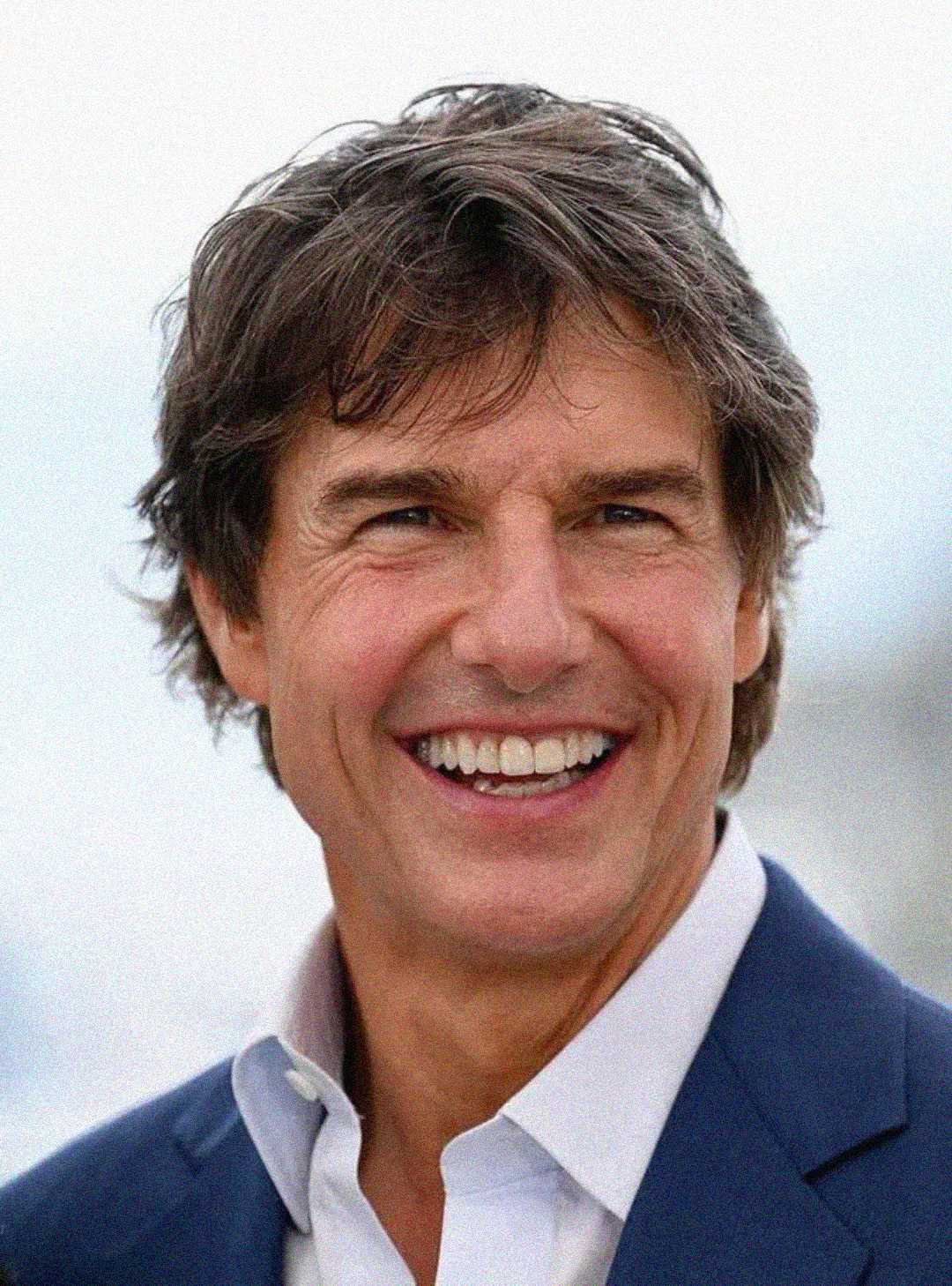 A man with tousled brown hair is smiling broadly, wearing a white collared shirt and a navy blue suit jacket. The background is softly blurred and light in color.