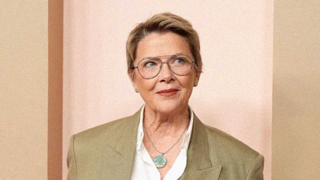 A person with short light brown hair and glasses, wearing a light brown jacket, white shirt, and a turquoise pendant necklace, stands in front of a pale pink background, looking slightly to the side and smiling softly.