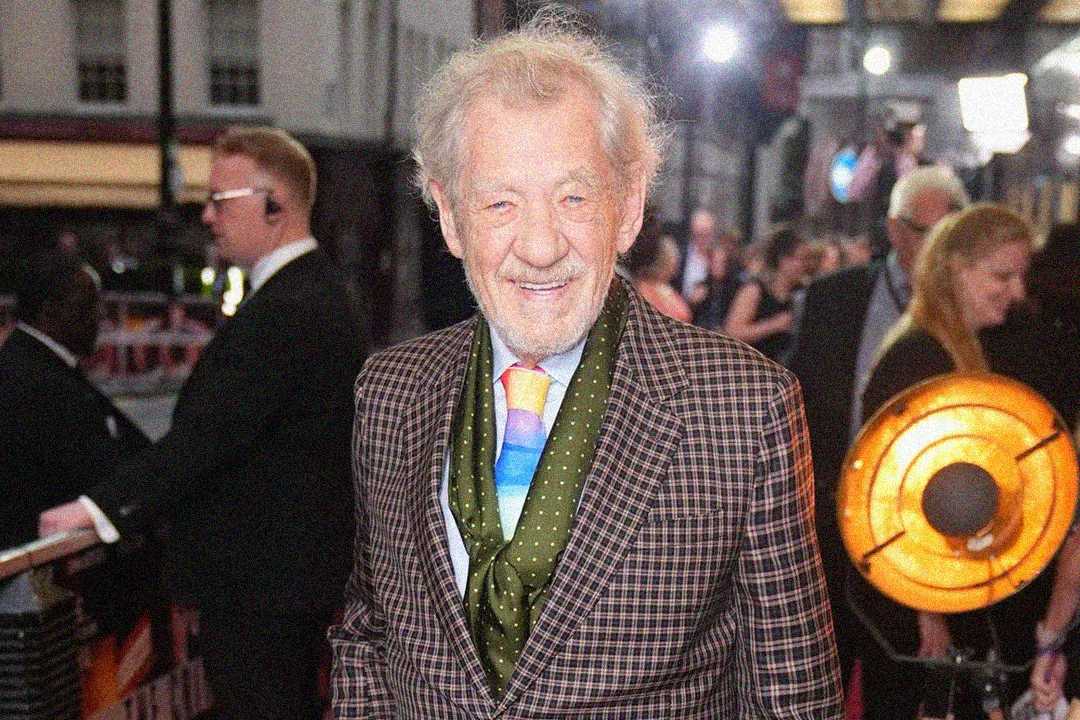 An older man in a checkered suit and colorful tie smiles at the camera on a red carpet, with people and lights in the background.