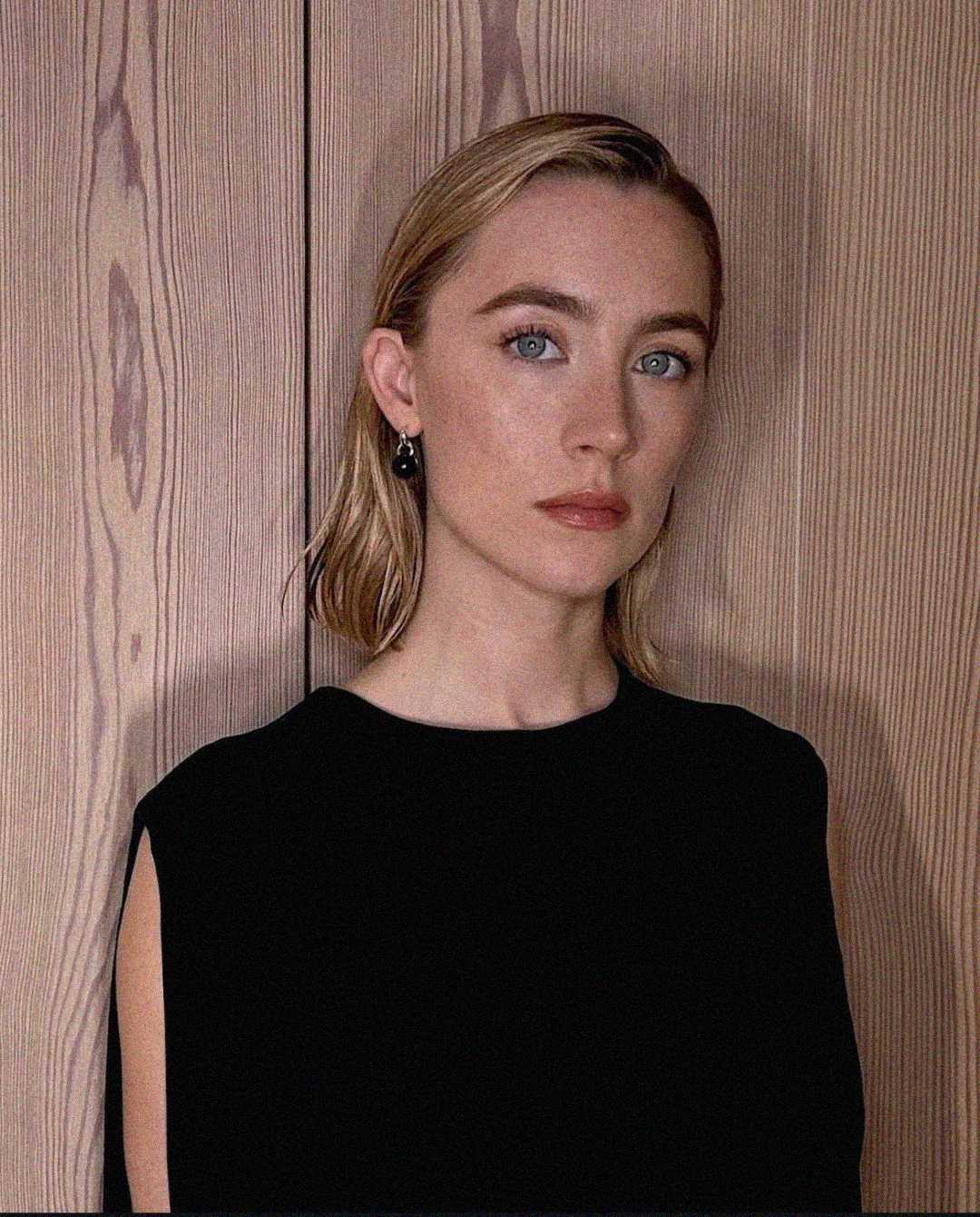 A woman with light skin, blonde hair, and blue eyes stands in front of a wooden wall. She is wearing a sleeveless black top and black earrings, and she has a neutral expression.