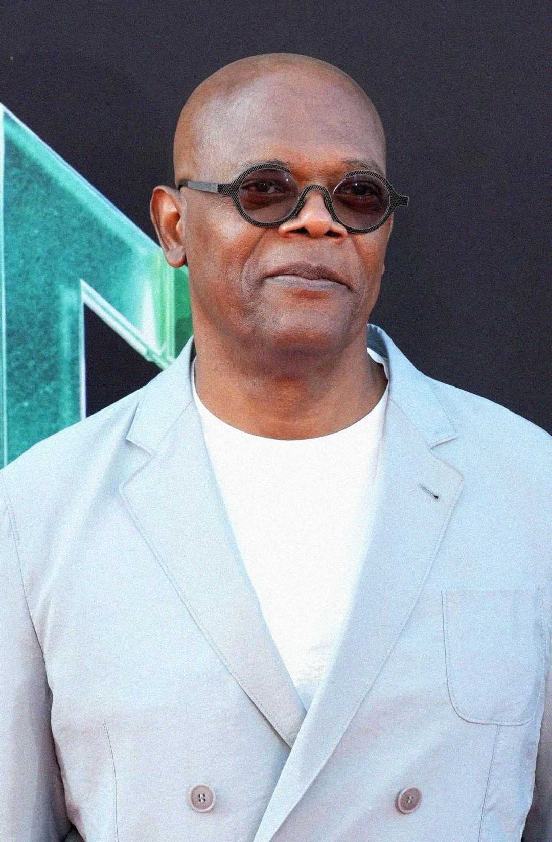 A man wearing round sunglasses, a light gray suit, and a white shirt stands in front of a dark background with a green geometric design.