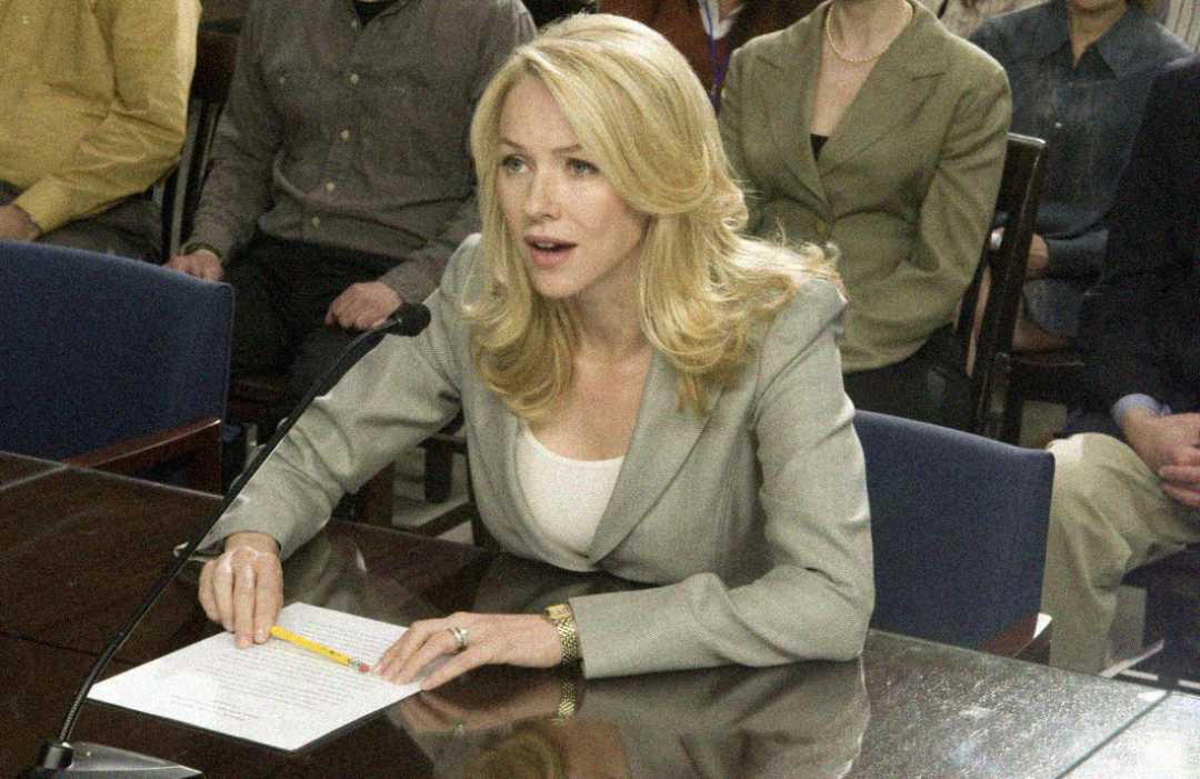 A woman with blonde hair in a gray blazer sits at a table with a microphone, holding papers, speaking during a formal meeting or hearing. Several people are seated in the background.