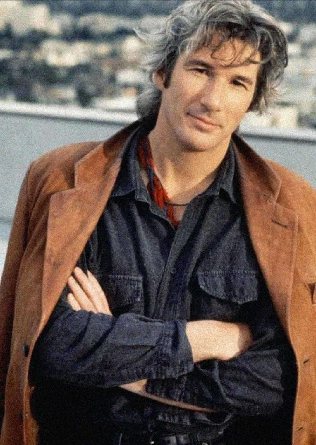 A man with wavy gray hair stands outdoors, arms crossed, wearing a brown suede jacket over a dark shirt and scarf, with a blurred cityscape in the background.