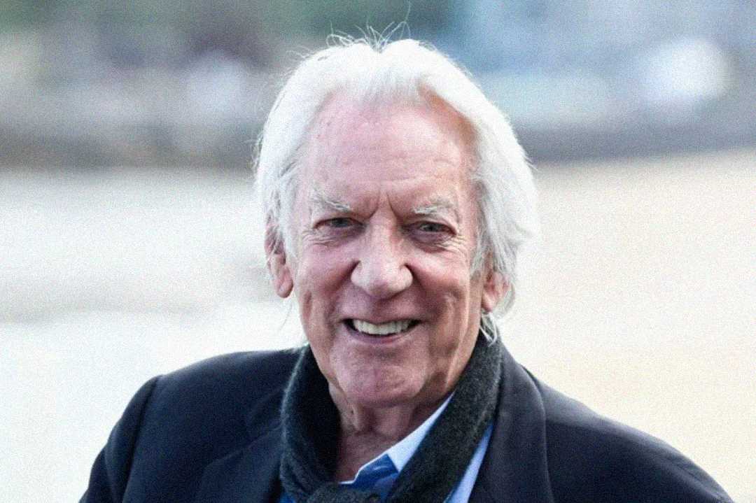 An elderly man with white hair is smiling outdoors, wearing a dark coat, scarf, and blue shirt. The background is blurred, suggesting a bright, open setting.