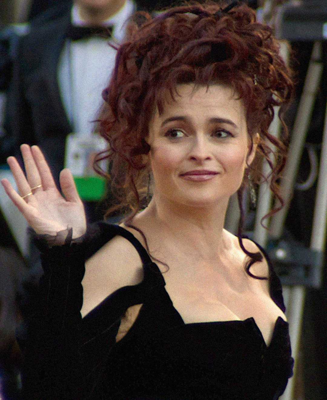 A woman with curly, dark hair in an updo, wearing a black dress, waves and smiles at an event. The background is blurred, showing people in formal attire and camera equipment.