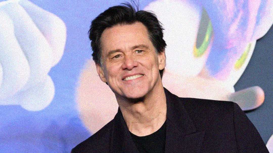 A man with dark hair in a dark blazer smiles at the camera, standing in front of a colorful animated background featuring a blue character with green eyes.