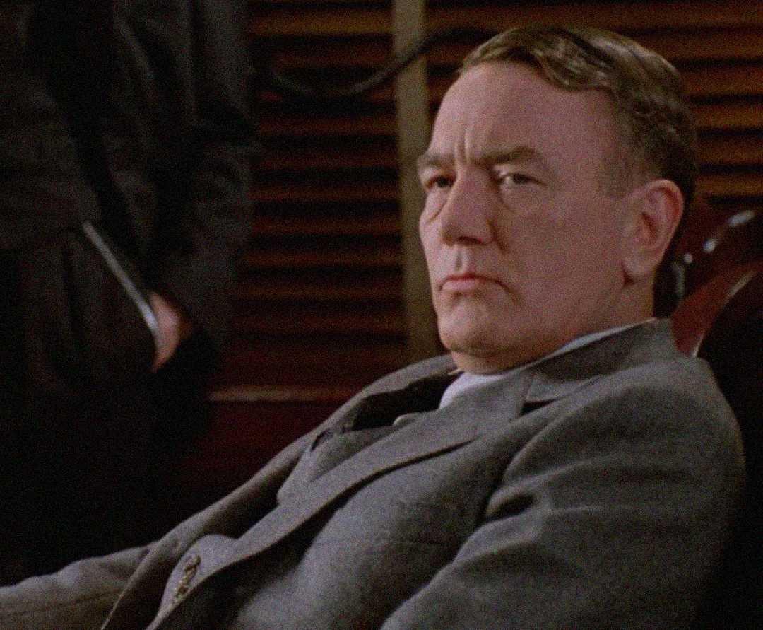 A man in a gray suit sits indoors, looking seriously to the side. His hair is neatly combed, and he appears to be in a formal setting with dark wood paneling in the background.