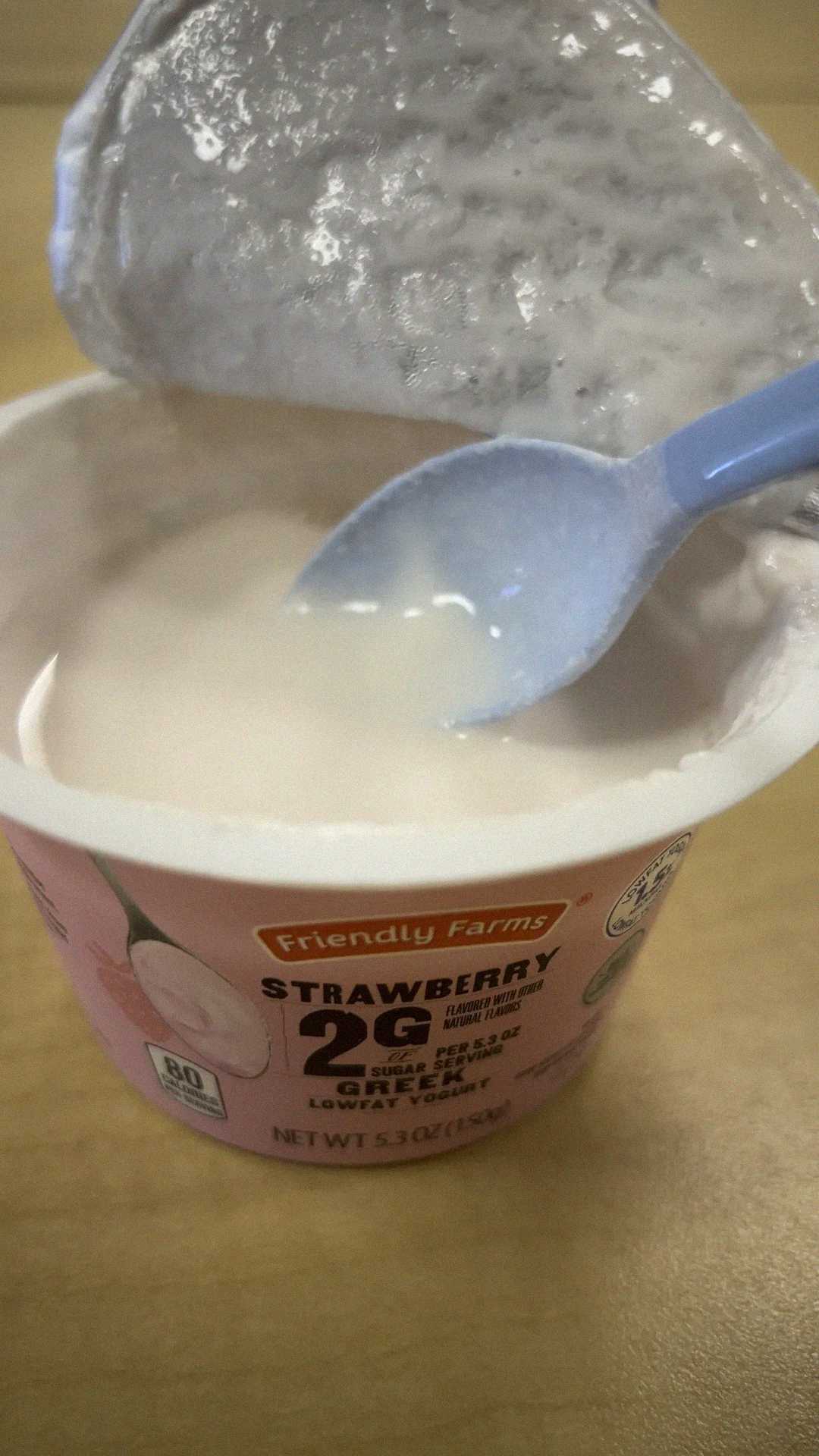 A partially opened container of Friendly Farms strawberry yogurt with a blue spoon inside, sitting on a light-colored surface. The foil lid is peeled back and some yogurt is on the spoon.