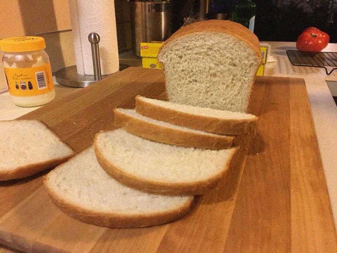 A loaf of white bread is partially sliced on a wooden cutting board, with several slices fanned out. Nearby are a jar, paper towel roll, and a tomato on a kitchen counter.