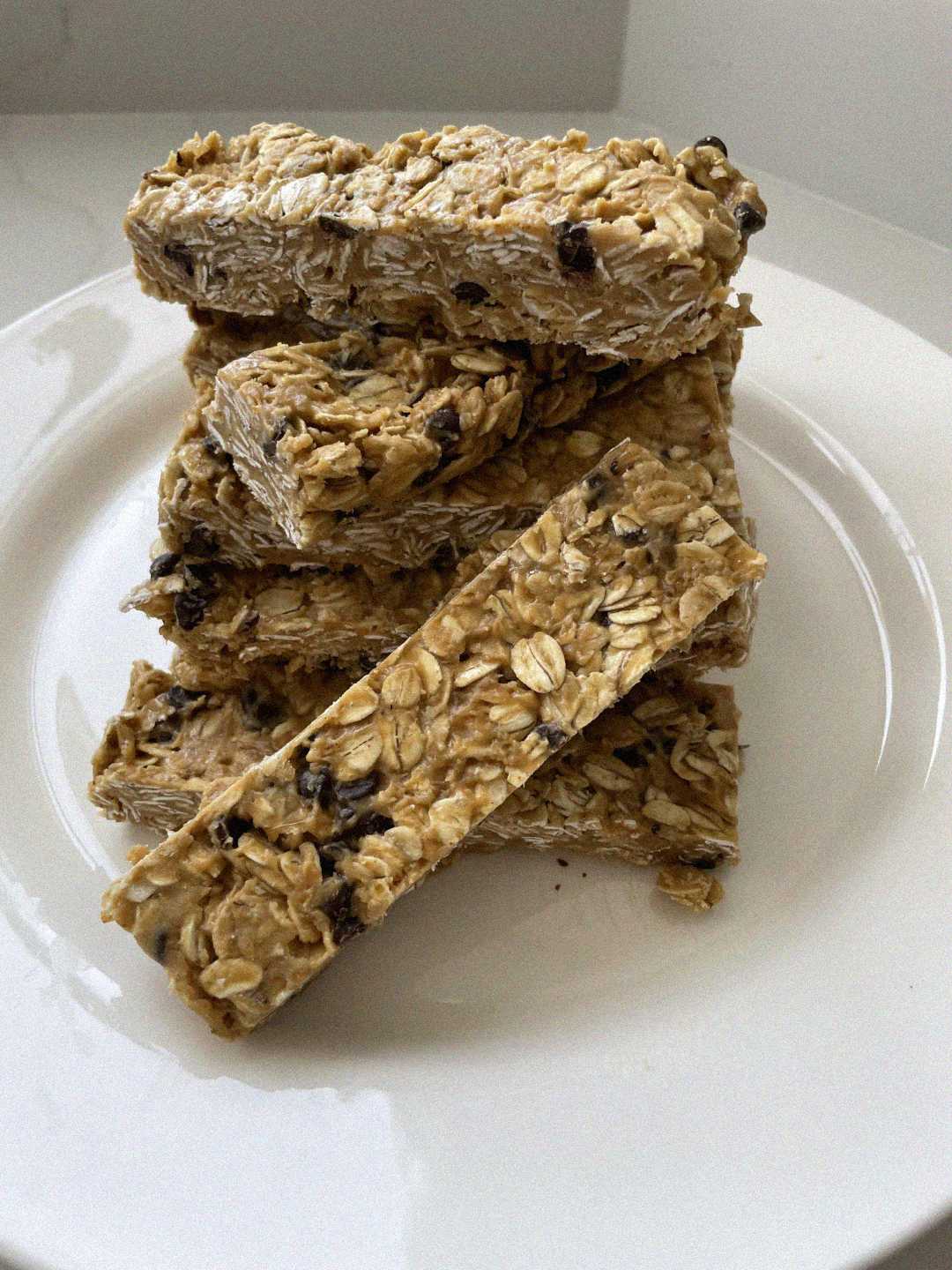 A stack of homemade oat granola bars with visible raisins is arranged on a white plate. The bars appear chewy and textured, set against a plain background.