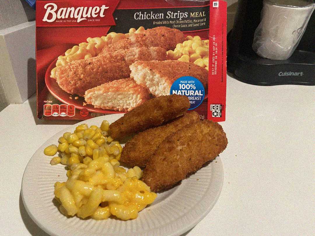 A plate with breaded chicken strips, macaroni and cheese, and corn sits in front of a Banquet Chicken Strips Meal box on a white countertop.