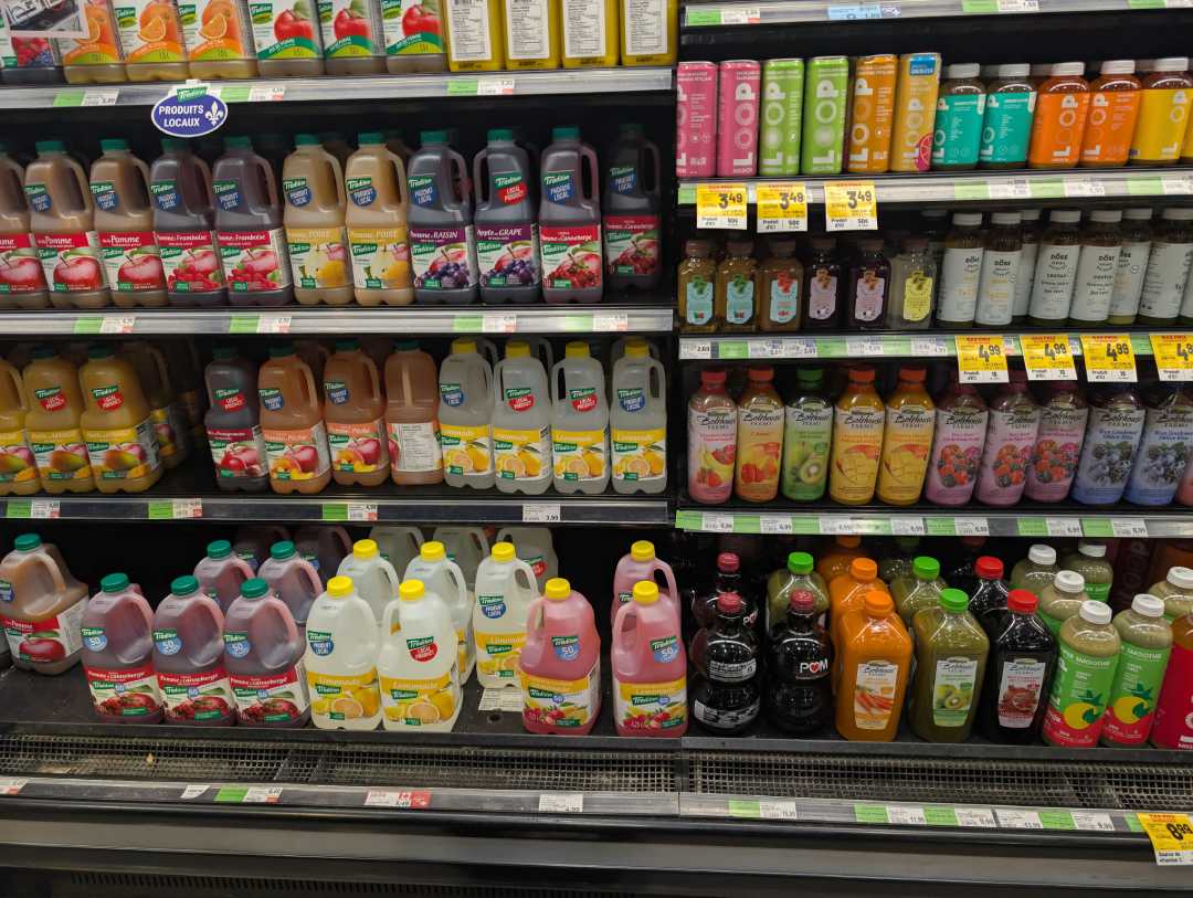 Refrigerated grocery store shelves filled with various bottled and jugged juices, including brands like Minute Maid and Tropicana. Price tags are visible, and the juices are in different colors and flavors.