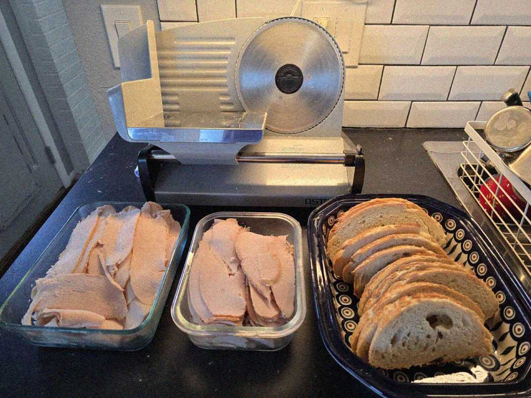 A meat slicer sits on a kitchen counter behind two glass containers of sliced deli meat and a ceramic dish holding several slices of bread. White subway tile backsplash is visible in the background.