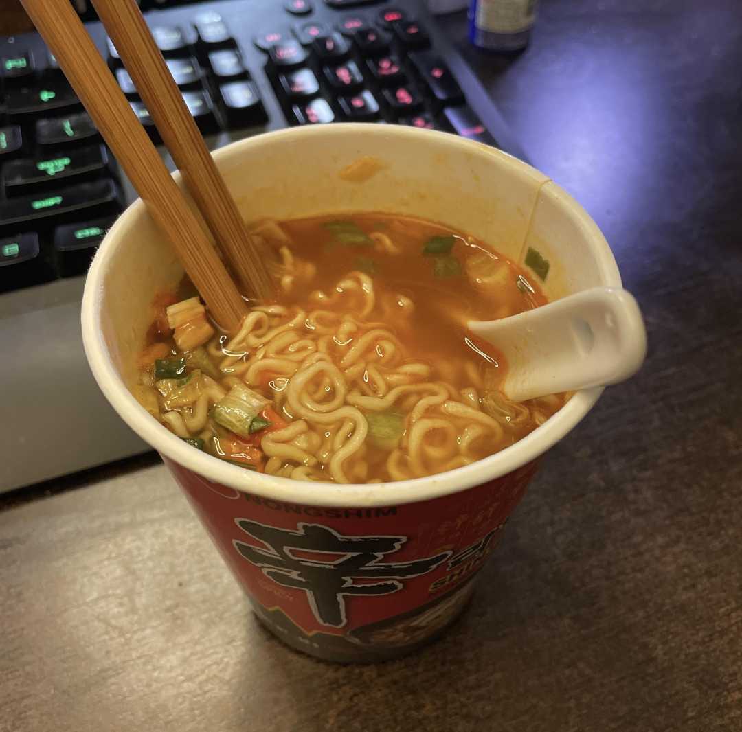 A cup of instant ramen noodles with chopsticks and a plastic spoon, placed on a wooden desk next to a keyboard. The soup appears hot with visible noodles and vegetables.