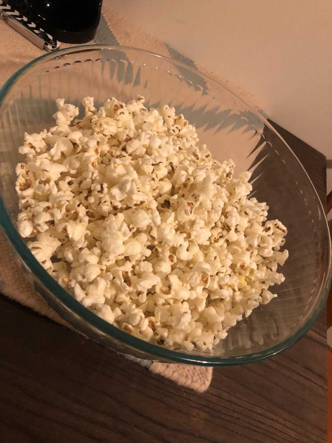A large glass bowl filled with popped popcorn sits on a dark wooden table, with a beige cloth underneath.
