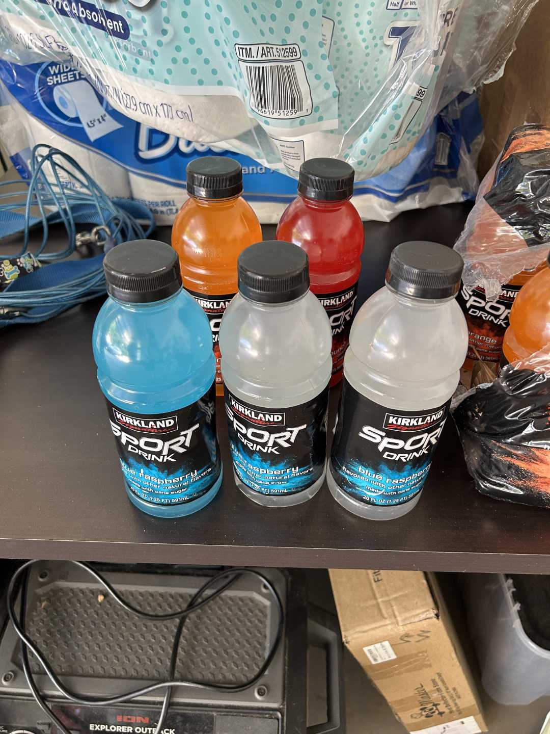 Six bottles of Kirkland Sport Drink in various flavors and colors are displayed on a shelf, with packs of bottled water and other miscellaneous items in the background.