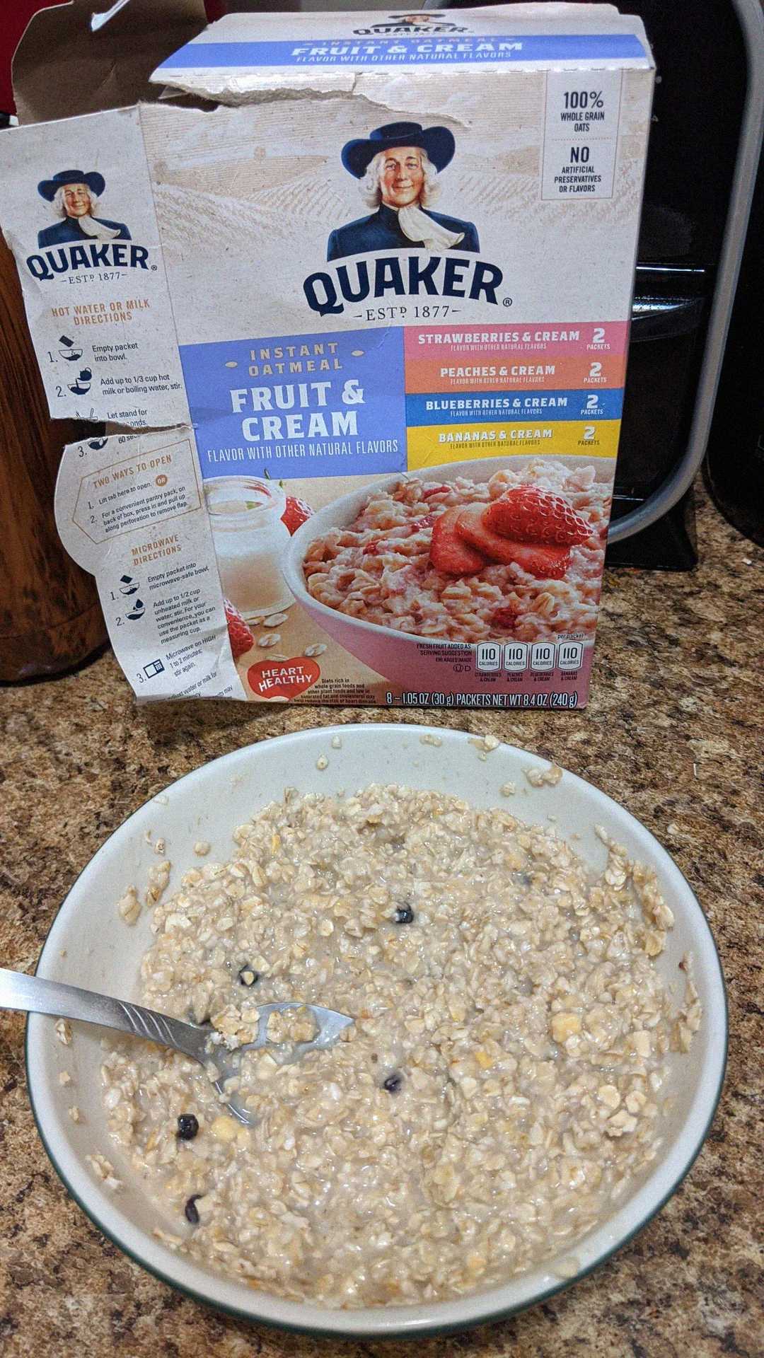 A bowl of oatmeal with visible blueberries sits on a counter in front of a Quaker Fruit & Cream instant oatmeal box. A spoon is in the bowl, and the box shows various oatmeal flavor options and nutritional info.