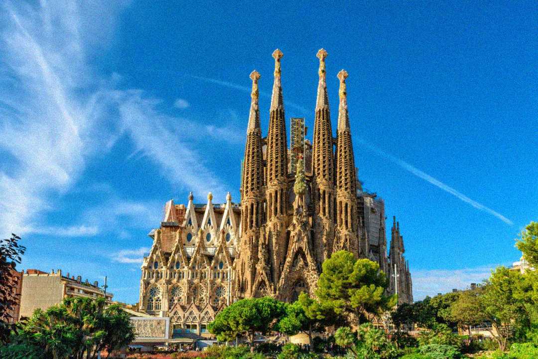 The Sagrada Familia basilica in Barcelona, Spain, with its tall spires and intricate façade, stands against a vivid blue sky, surrounded by green trees and city buildings.