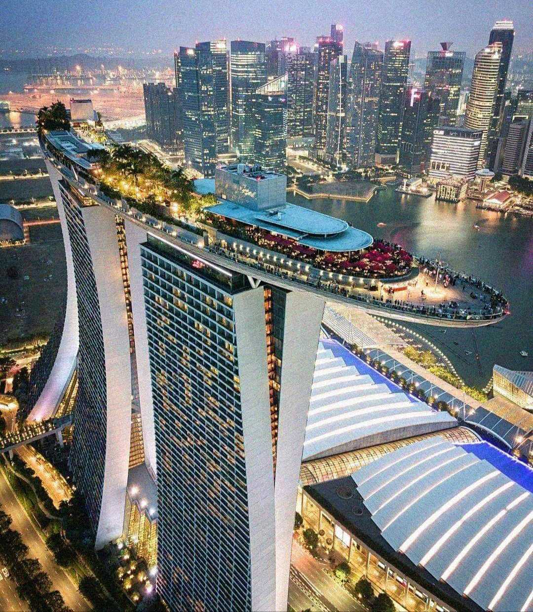 Aerial view of Marina Bay Sands in Singapore at dusk, showing its three towers connected by a rooftop SkyPark with a pool, surrounded by city skyscrapers and waterfront buildings.