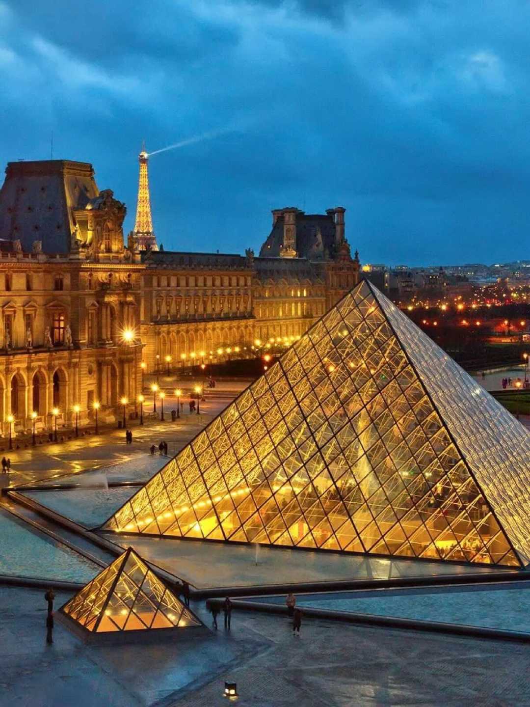 The illuminated glass pyramid of the Louvre Museum in Paris at dusk, with the Eiffel Tower glowing in the background and historic buildings lit up under a deep blue sky.