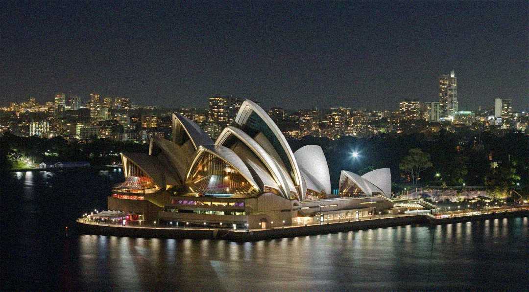 Sydney Opera House illuminated at night, surrounded by water, with city buildings and skyscrapers lit up in the background under a dark sky.