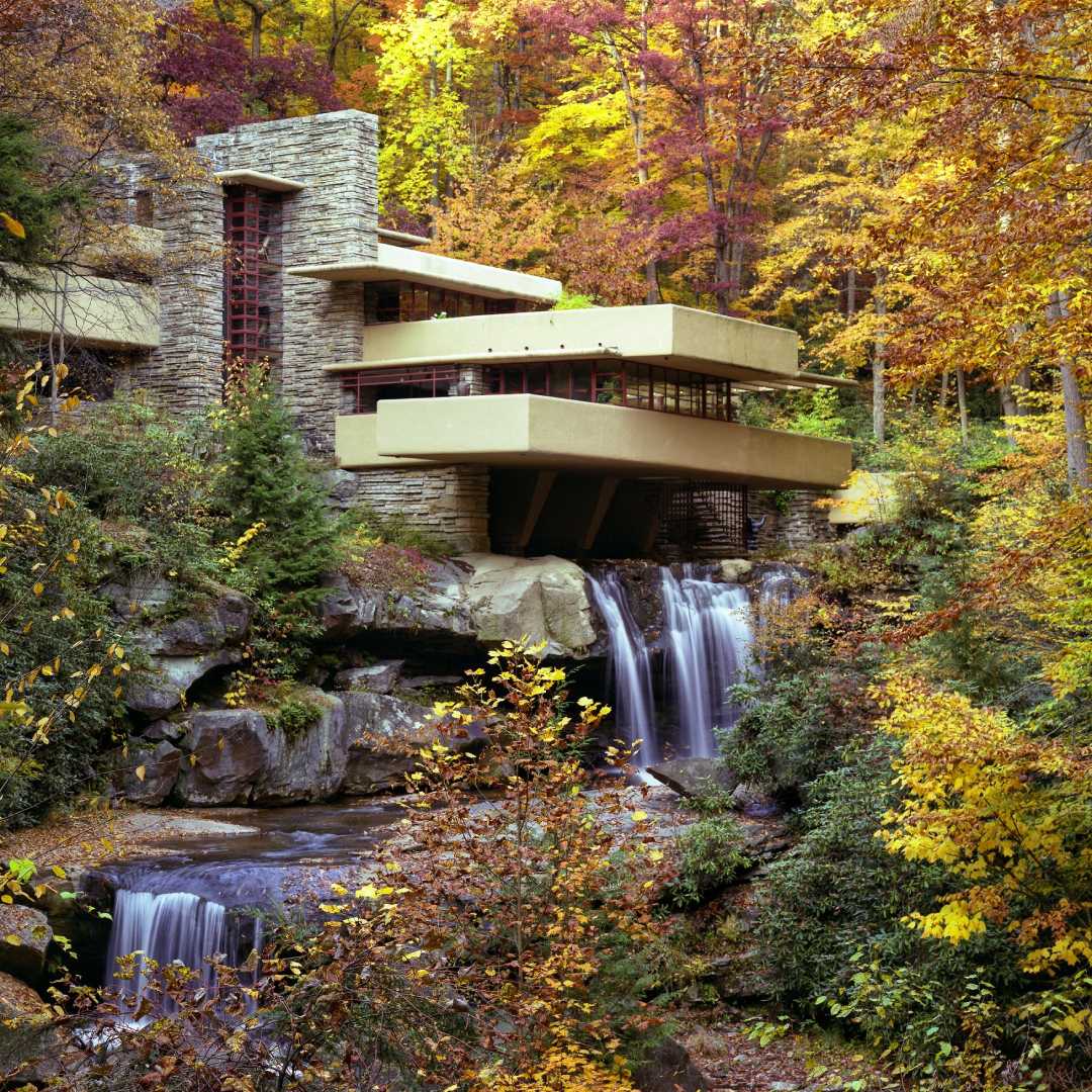 A modern house with stone and concrete terraces is built over a waterfall, surrounded by vibrant autumn trees in a forest. Water cascades below, and colorful leaves frame the peaceful scene.