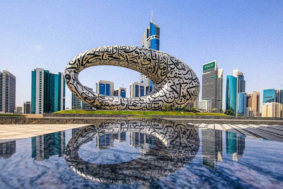 A futuristic torus-shaped building with Arabic calligraphy on its surface stands among modern skyscrapers, with its reflection visible in a pool of water in the foreground. The sky is clear and blue.