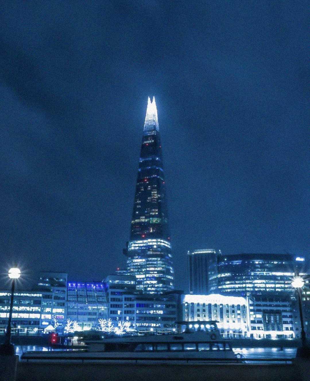 The Shard skyscraper in London illuminated at night, with its pointed top glowing against a dark, cloudy blue sky. Surrounding buildings and streetlights are also lit, reflecting in the foreground.