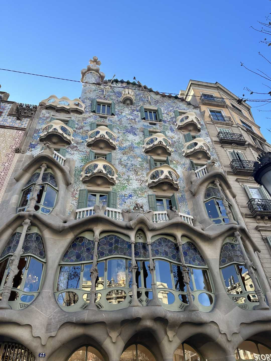 A view of Casa Batlló in Barcelona, featuring its colorful mosaic façade, organic-shaped balconies, and wavy architectural lines under a clear blue sky.