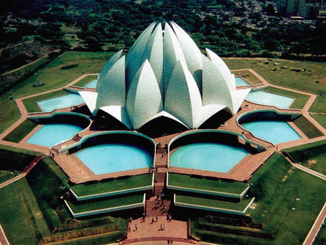 Aerial view of the Lotus Temple in Delhi, India, featuring white petal-like structures surrounded by blue pools and green landscaped gardens.