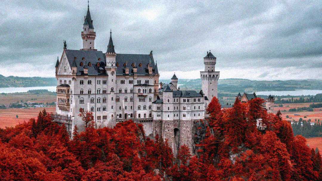 A large, fairytale-like castle with tall towers sits atop a rocky hill, surrounded by vibrant red autumn trees, under a cloudy sky with fields and distant water in the background.