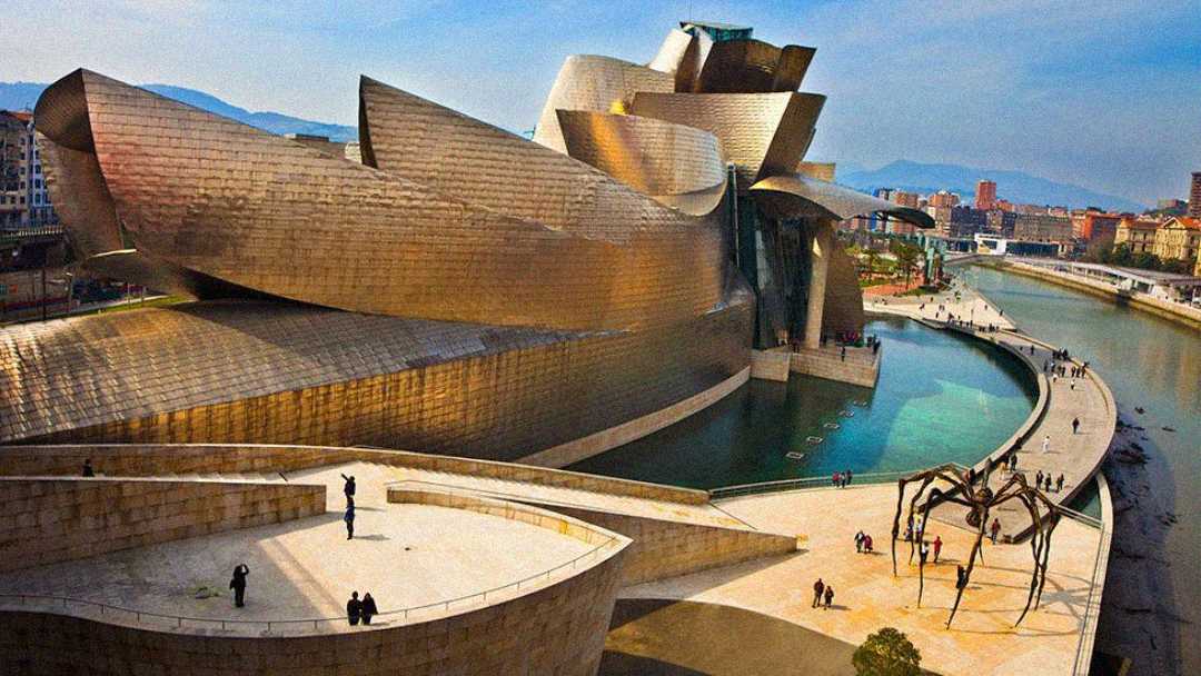 The Guggenheim Museum Bilbao, a striking modern building with curving, metallic surfaces, sits beside a river. Nearby is a large spider sculpture and people walking along the river promenade. Mountains rise in the background.
