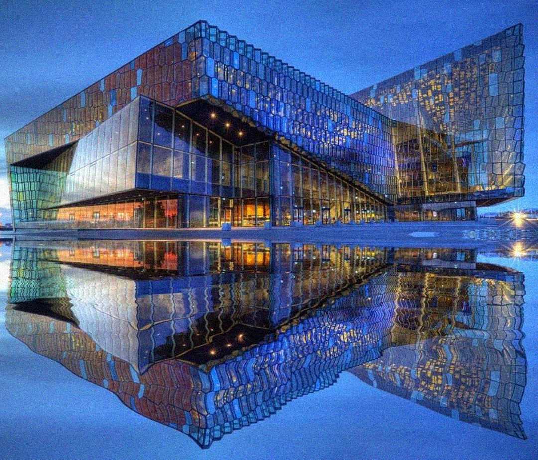 A modern glass building with colorful, geometric panels is dramatically lit at dusk and reflected perfectly in a smooth body of water below.