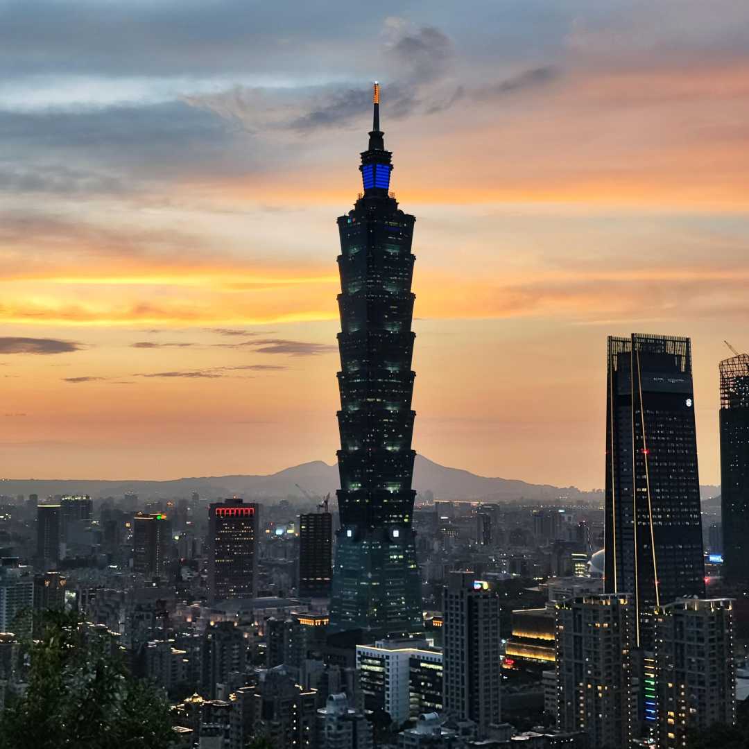 Taipei 101 skyscraper rises above the city skyline at sunset, with a warm, colorful sky and scattered clouds in the background, surrounded by other buildings in Taipei, Taiwan.