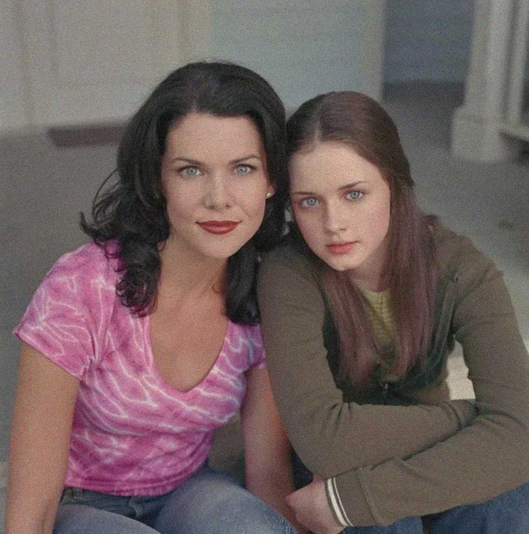 Two women with dark hair sit closely together, looking at the camera. One wears a pink tie-dye shirt, the other a green jacket. They appear to be indoors, sitting on a carpeted floor near a doorway.