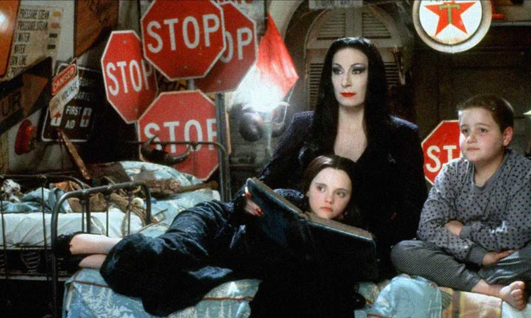 A woman in black sits on a bed reading to two children. The room is cluttered with multiple stop signs on the wall and various vintage items. The girl lies on the woman's lap while the boy sits beside them, smiling.