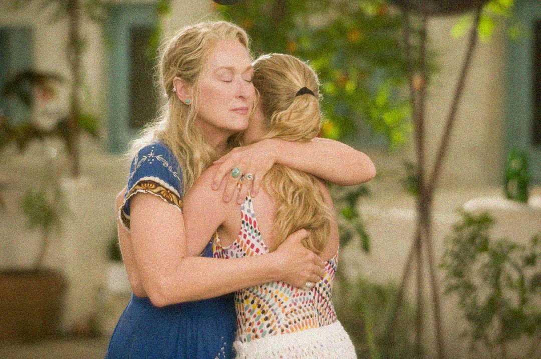 Two women with long blonde hair warmly embrace each other outdoors, one in a blue dress and the other in a colorful patterned outfit, with soft sunlight and greenery in the background.